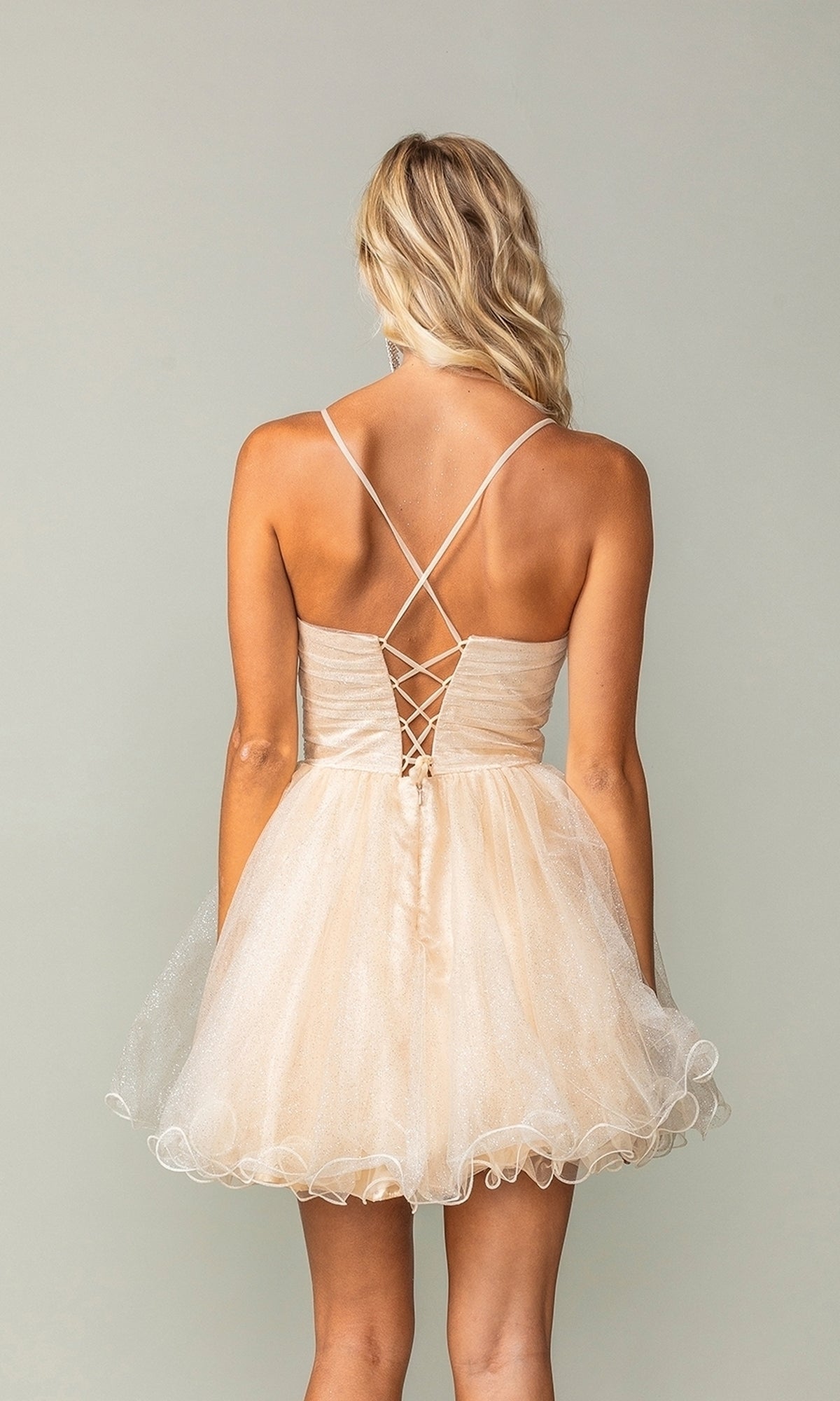 A woman in the Lace-Up Short Babydoll Homecoming Dress 3385—light beige, sleeveless, backless with crisscross straps and a ruffled short skirt—stands against a plain background.